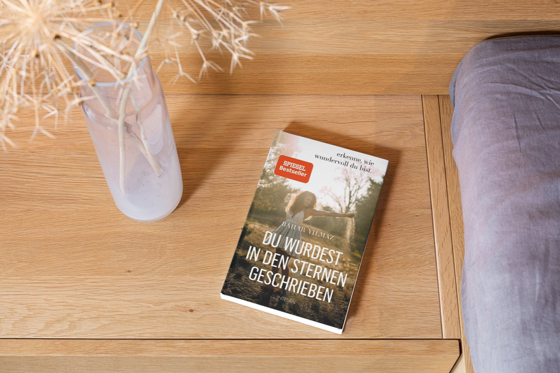 Ein Buch mit dem Titel „Du wurdest in den Sternen geschrieben“ liegt auf einer Holzfläche neben einer hellen Vase mit getrockneten Blumen. Rechts ist der Teil eines grauen Kissens zu sehen.