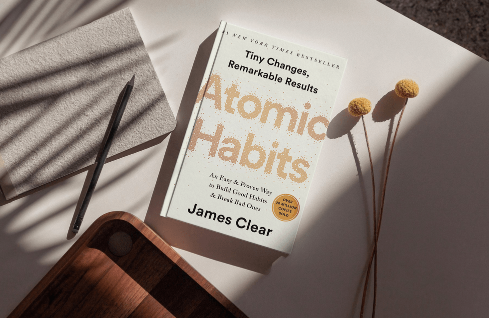 Ein Exemplar von „Atomic Habits“ von James Clear liegt auf einem Tisch. Ein Bleistift und zwei gelbe Blumen im Schatten begleiten es. Der Untertitel des Buches lautet: „Ein einfacher und bewährter Weg, gute Gewohnheiten aufzubauen und schlechte zu vermeiden.“.