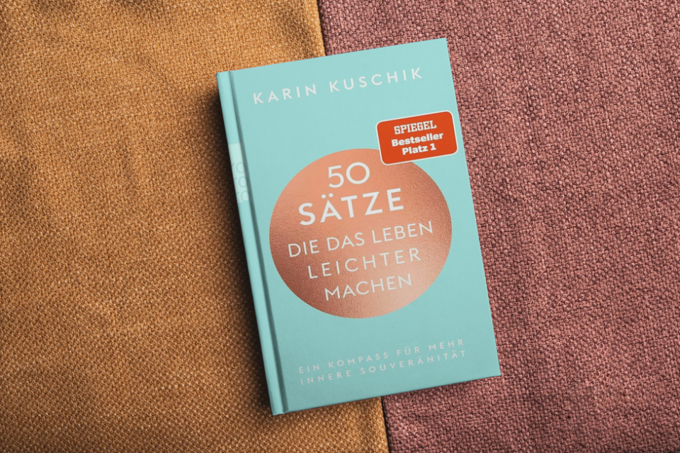 Auf einer strukturierten Stoffoberfläche in Braun- und Rosatönen liegt ein türkisfarbenes Buch mit dem Titel „50 Sätze die das Leben leichter machen“ von Karin Kuschik, versehen mit einem roten „Spiegel Bestseller Platz 1“-Aufkleber.