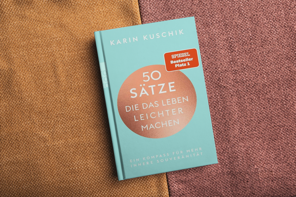 Auf einer strukturierten Stoffoberfläche in Braun- und Rosatönen liegt ein türkisfarbenes Buch mit dem Titel „50 Sätze die das Leben leichter machen“ von Karin Kuschik, versehen mit einem roten „Spiegel Bestseller Platz 1“-Aufkleber.