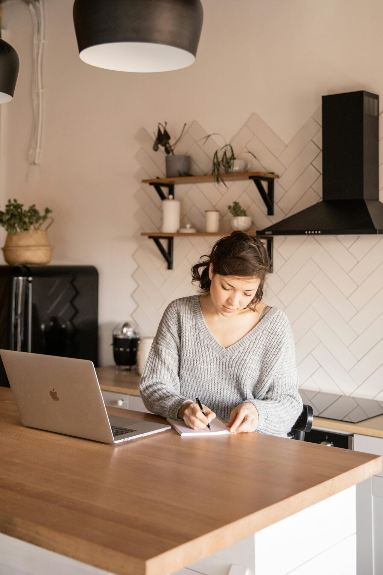 Frau plant Arbeit in den USA und schreibt mit einem Laptop in ein Notizbuch an ihrer Küchentheke.