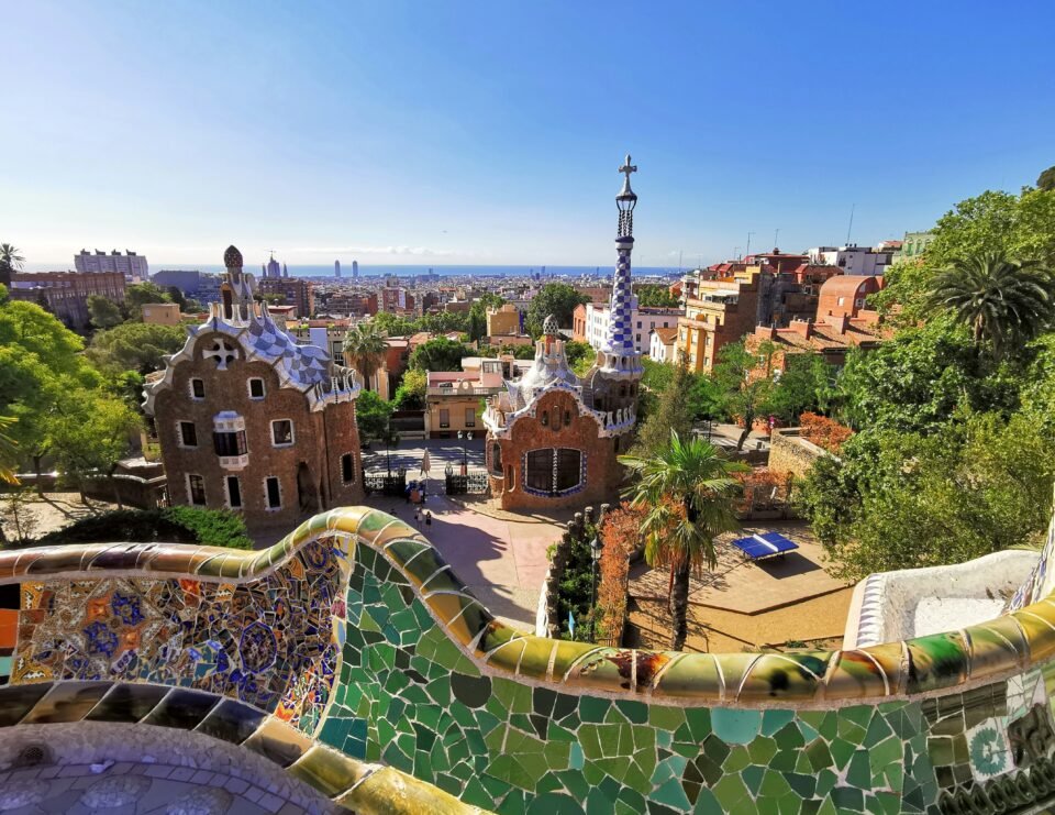 Bunte Mosaikbank mit Blick auf skurrile Geb&auml;ude im Park G&uuml;ell, Barcelona, an einem sonnigen Tag.