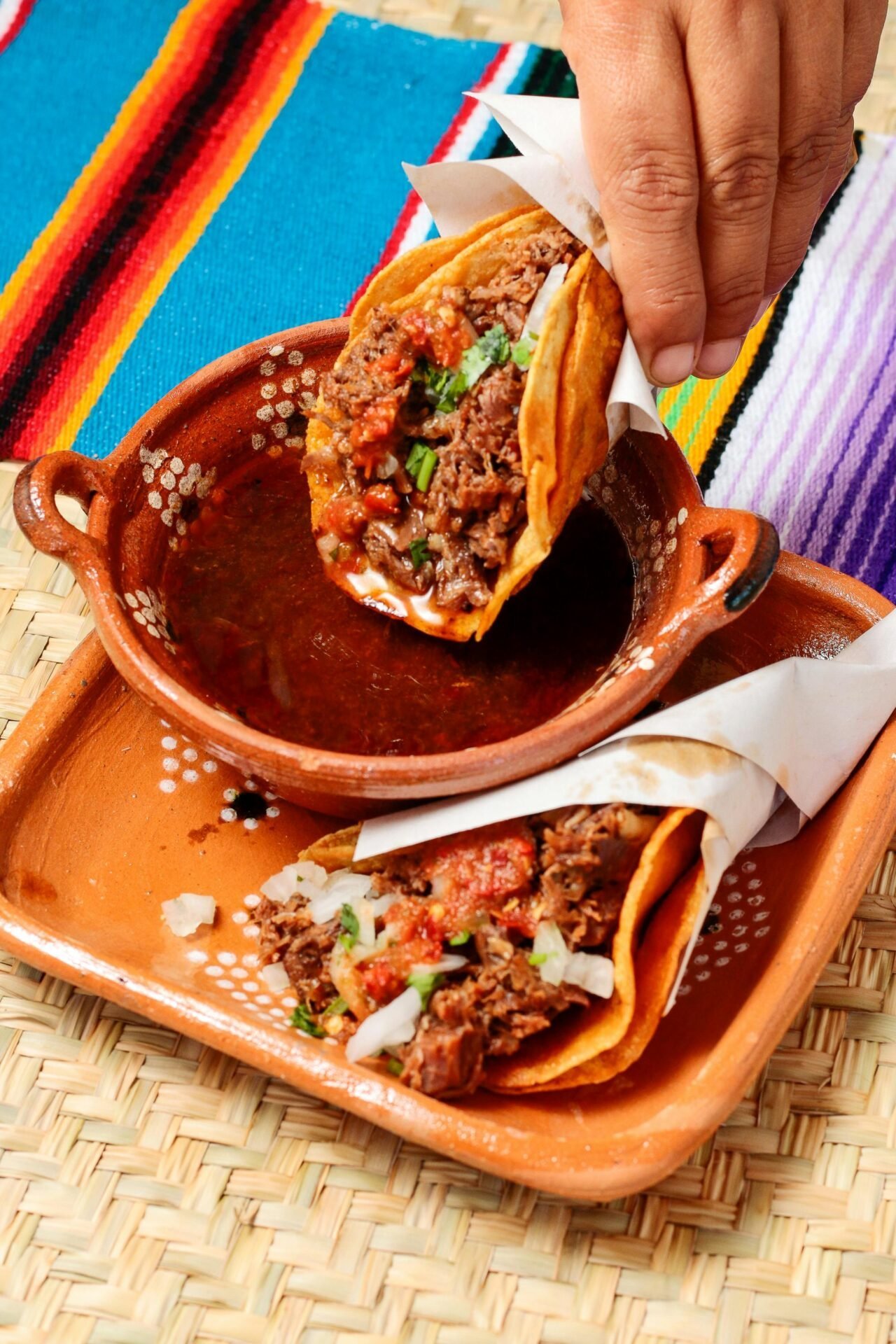 Tauchen Sie einen Birria-Taco mit der Hand in Consommé – Ihr Sommergenuss auf einer bunten Tischdecke.