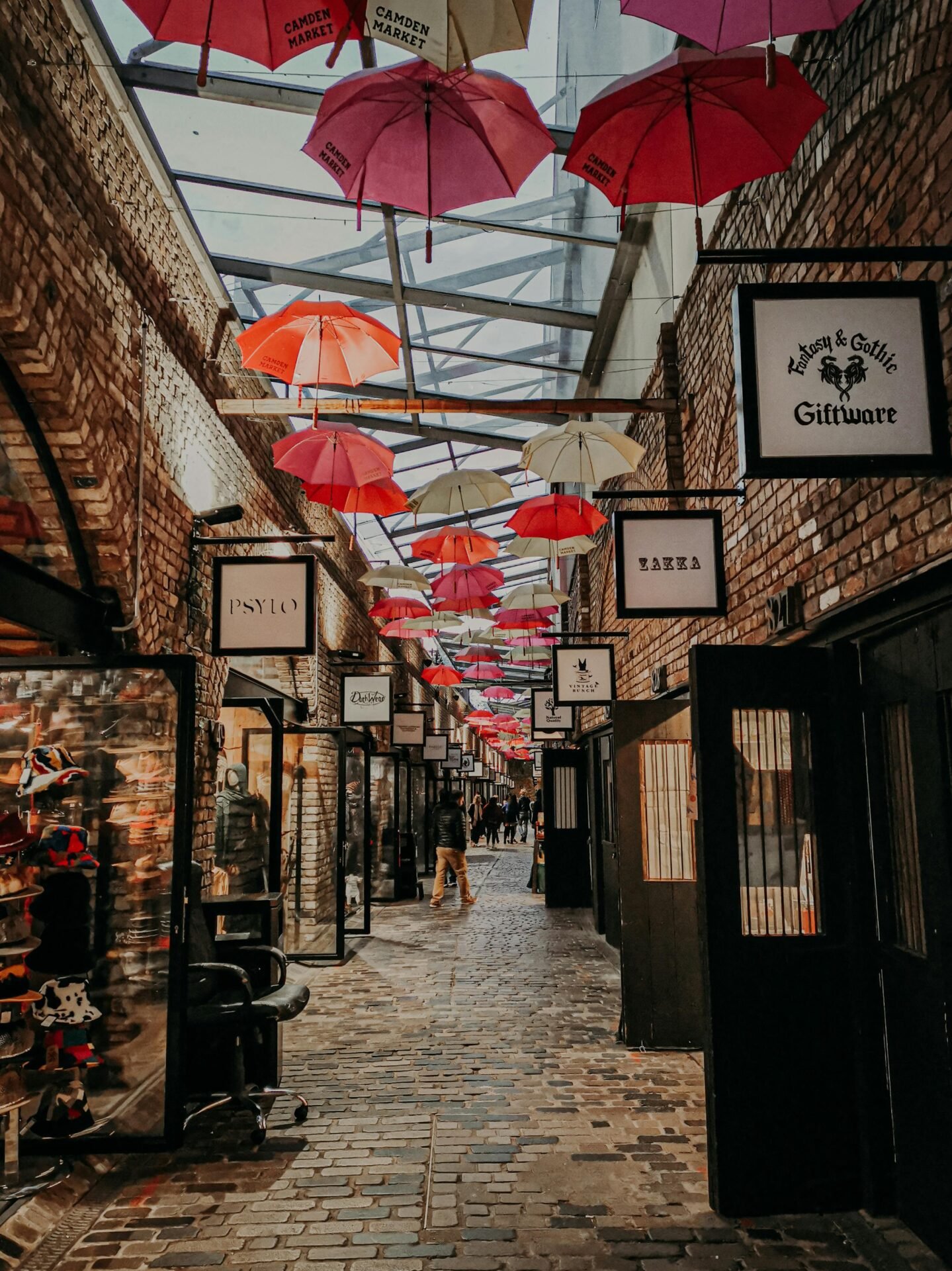 Backsteingasse in London mit hängenden roten und rosa Regenschirmen, gesäumt von Ladenschildern und offenen Türen.
