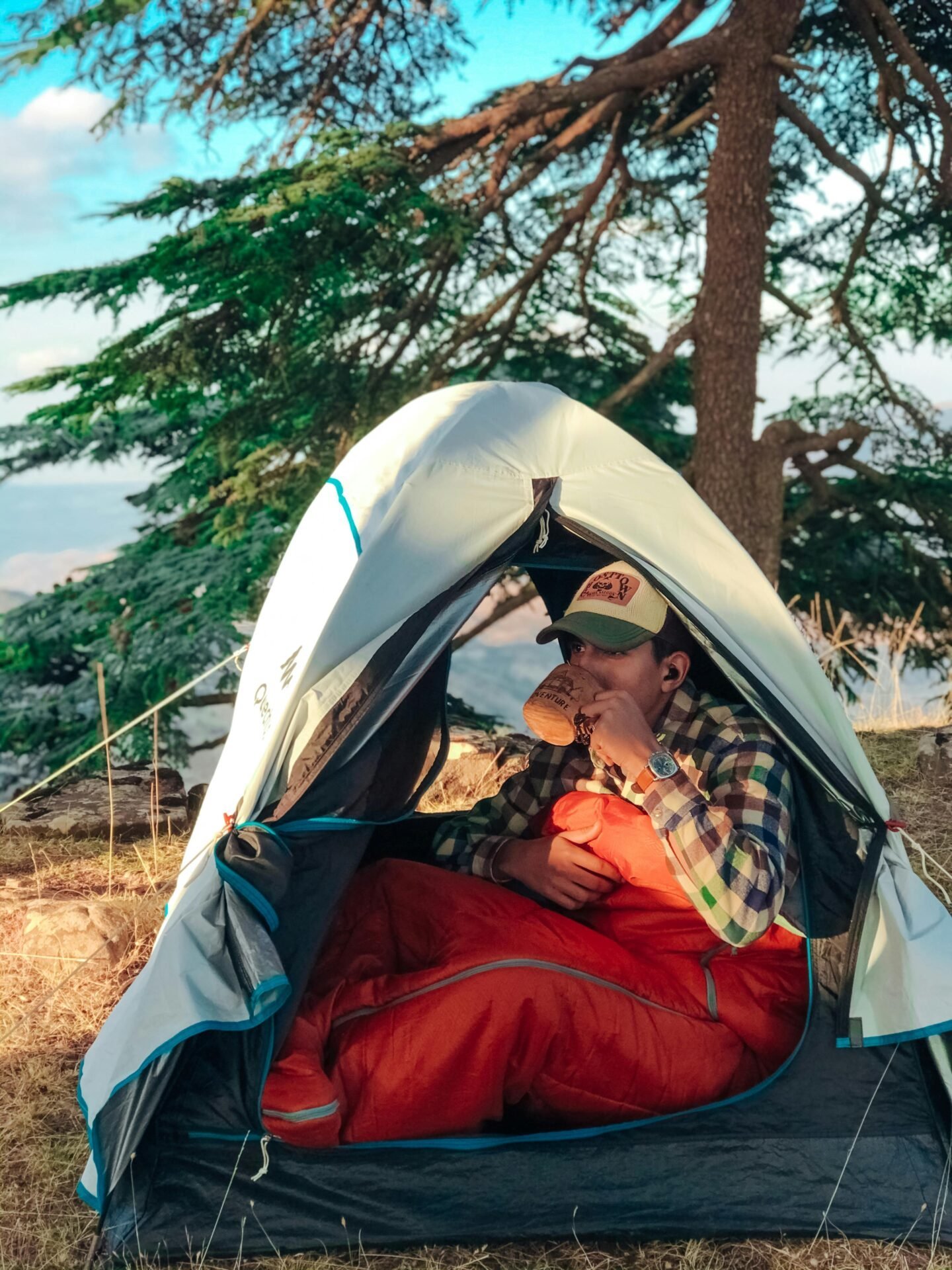 Eine Person in einem Zelt trinkt mithilfe von Camping-Hacks aus einem Becher, der in einen orangefarbenen Schlafsack eingewickelt ist, im Freien.