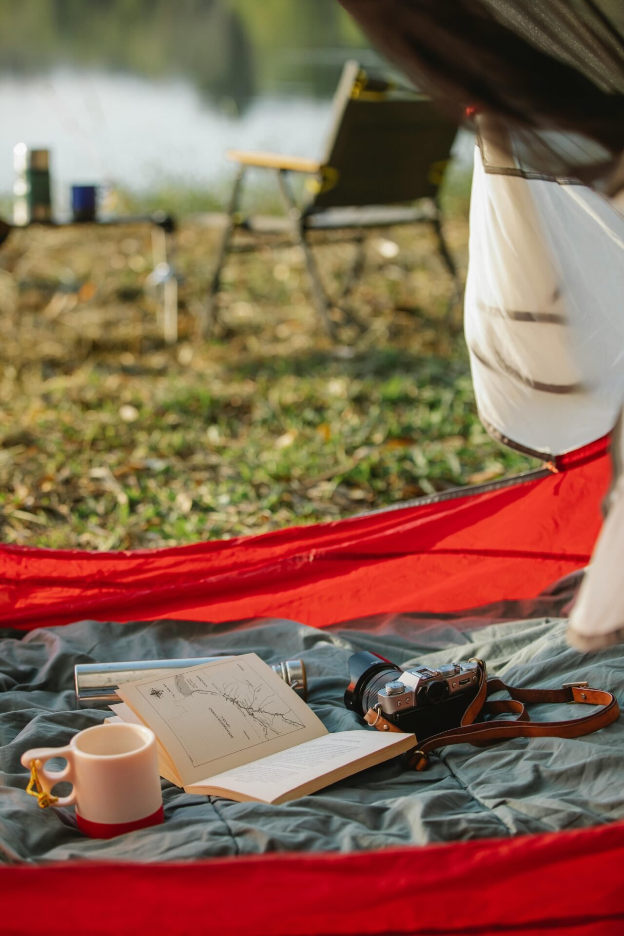 Offenes Zelt mit Campingausrüstung – Karte, Kamera, Tasse – auf einer Decke; dahinter Campingplatz und Stuhl.