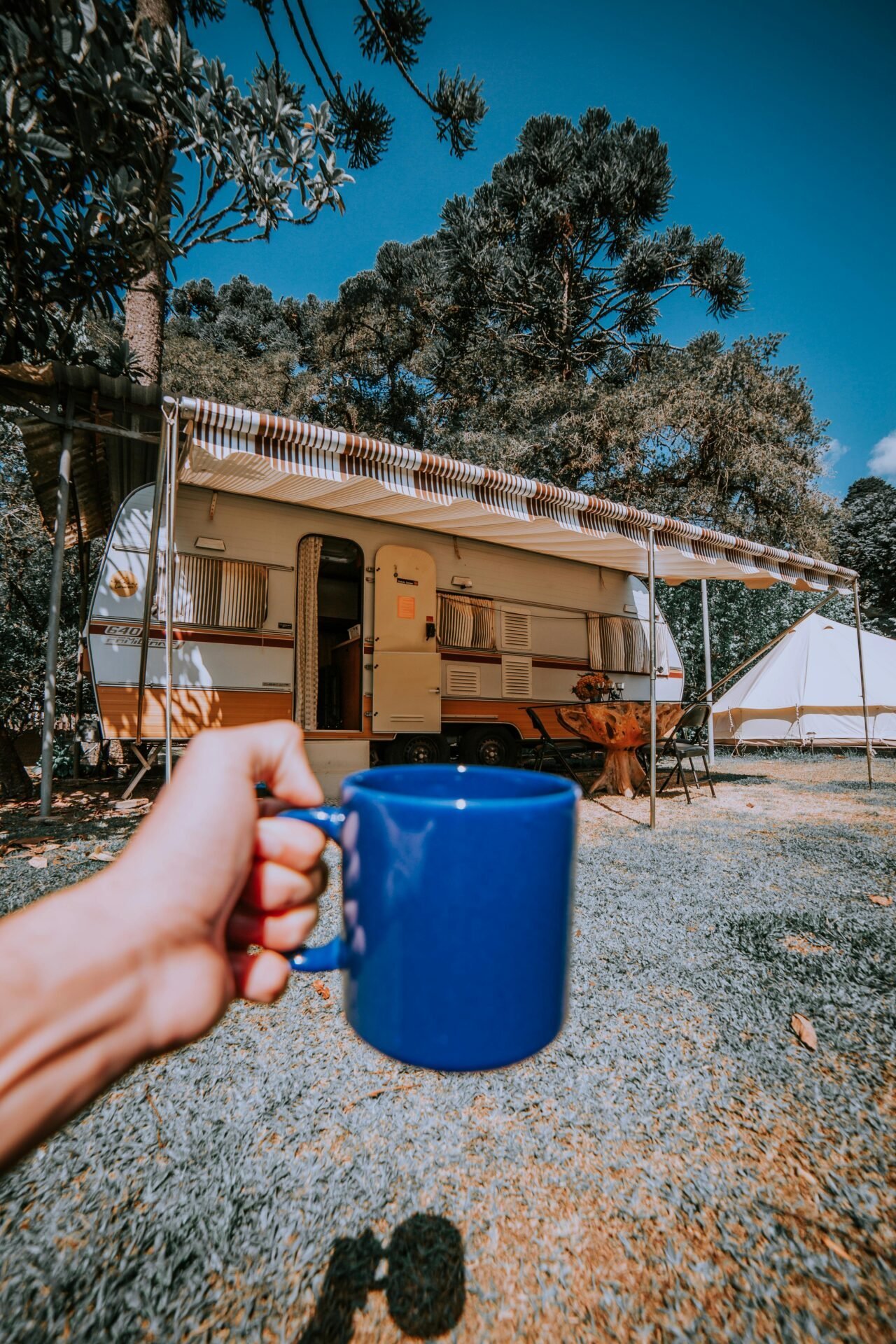 Eine Hand hält einen blauen Becher und genießt Campingsprüche neben einem Wohnmobil zwischen den Bäumen.