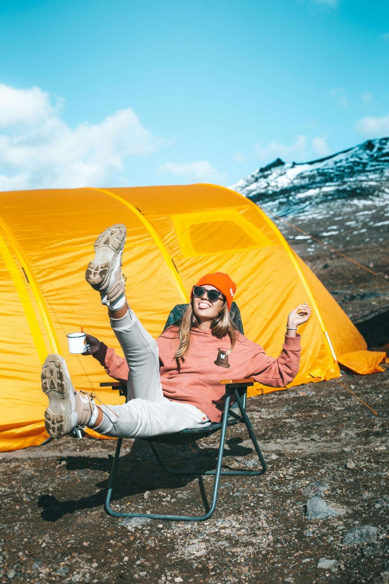 Frau mit orangefarbenem Hut entspannt sich neben einem gelben Zelt und hält eine Tasse – perfekt für Camping-Momente.