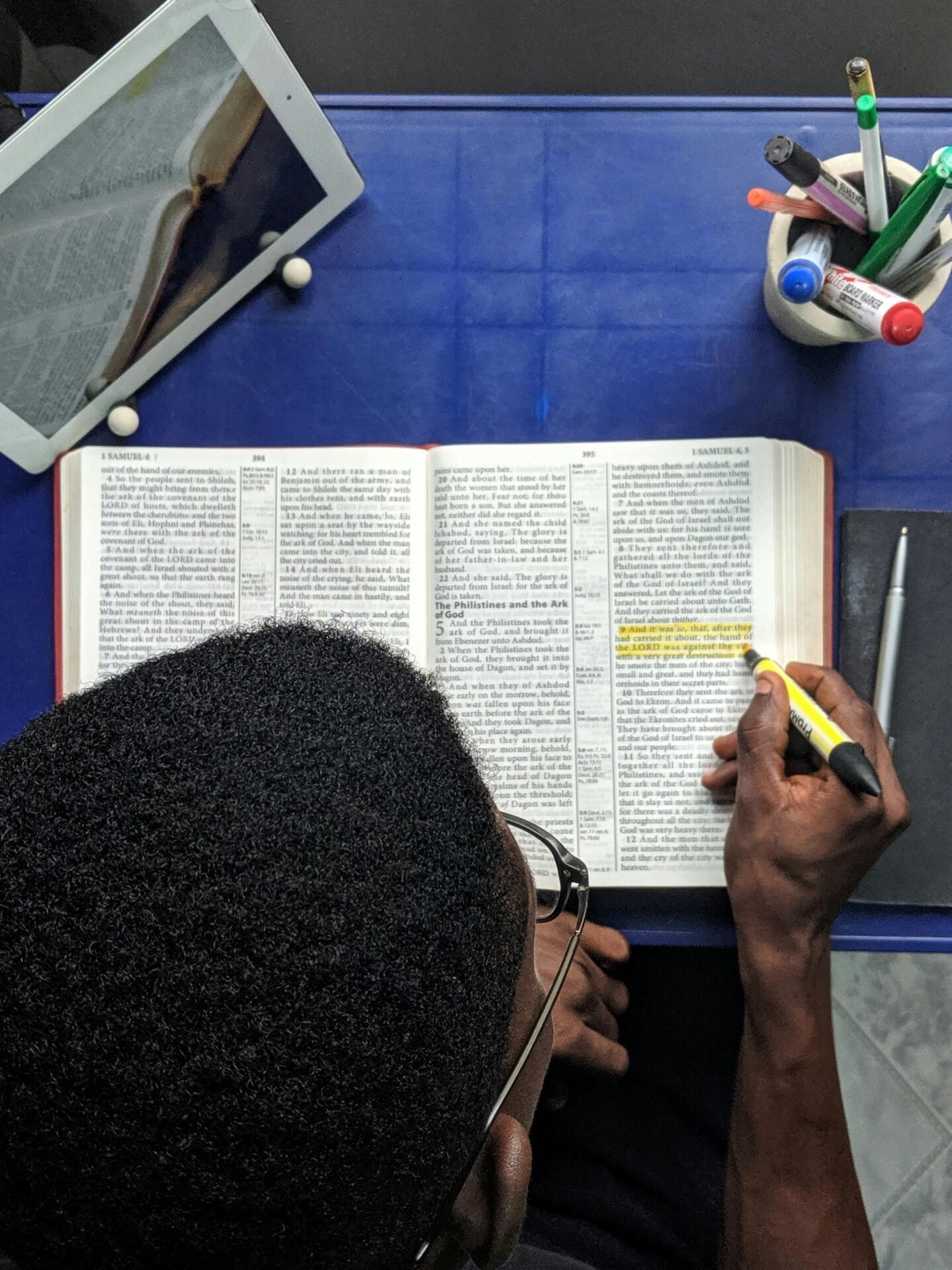 Person hebt Text im Buch hervor – erfolgreich lernen mit Tablet und Schreibwaren am blauen Tisch.