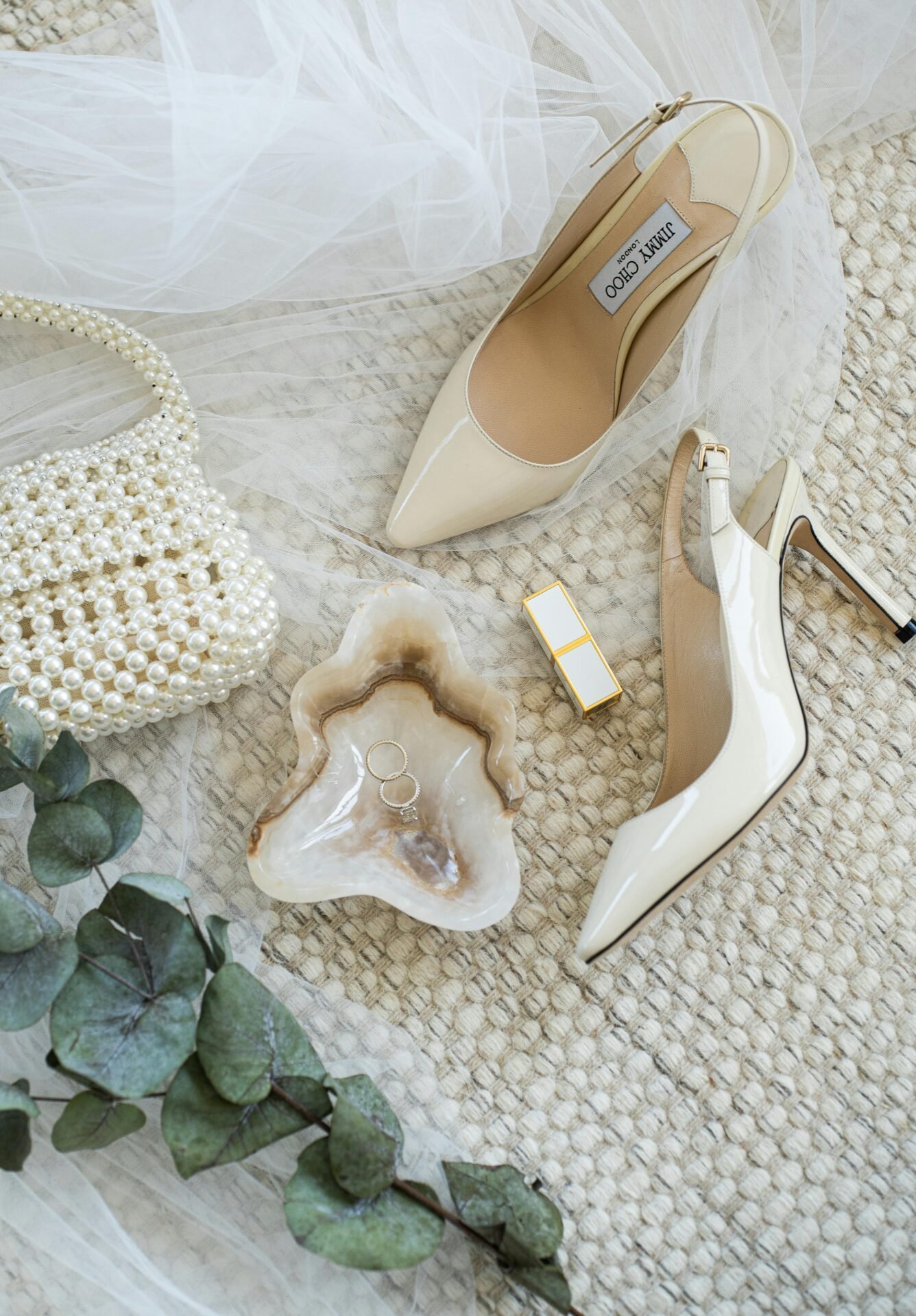 Cremefarbene Pumps, Perlenhandtasche, Lippenstift, Eheringe und Eukalyptus auf strukturiertem Stoff.
