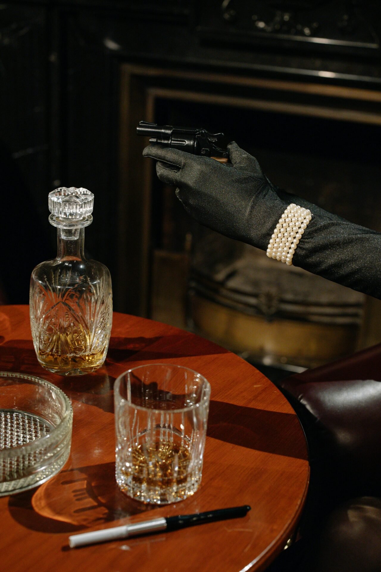 Eine behandschuhte Hand mit Perlen – erfolgreiche Frauen – hält eine Pistole neben Whisky und einem Aschenbecher.
