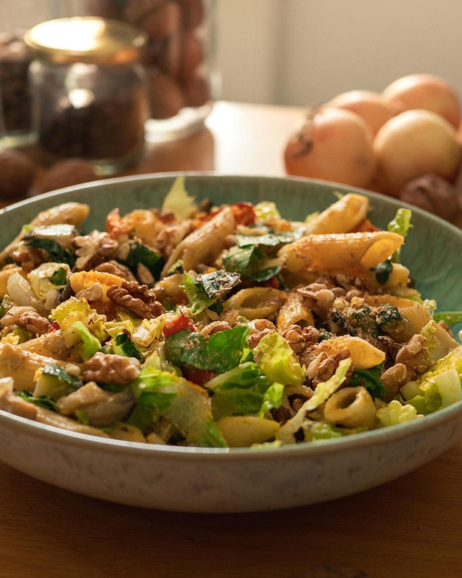 Eine Schüssel Penne-Nudelsalat – perfekt zum Essen bei der Arbeit – steht mit Zwiebeln und Gläsern im Hintergrund.