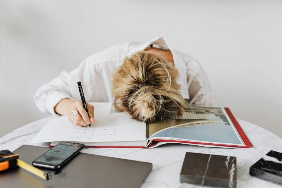 Person, die Familie und Karriere unter einen Hut bringen muss, ruht mit dem Kopf auf einem Buch inmitten von Arbeitsmaterialien auf einem Tisch.