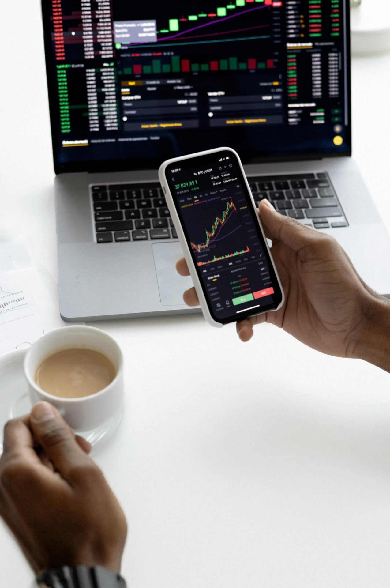 Person verfolgt finanzielle Freiheit am Telefon mit Aktienchart, Laptop-Grafiken und Kaffee in der Nähe.