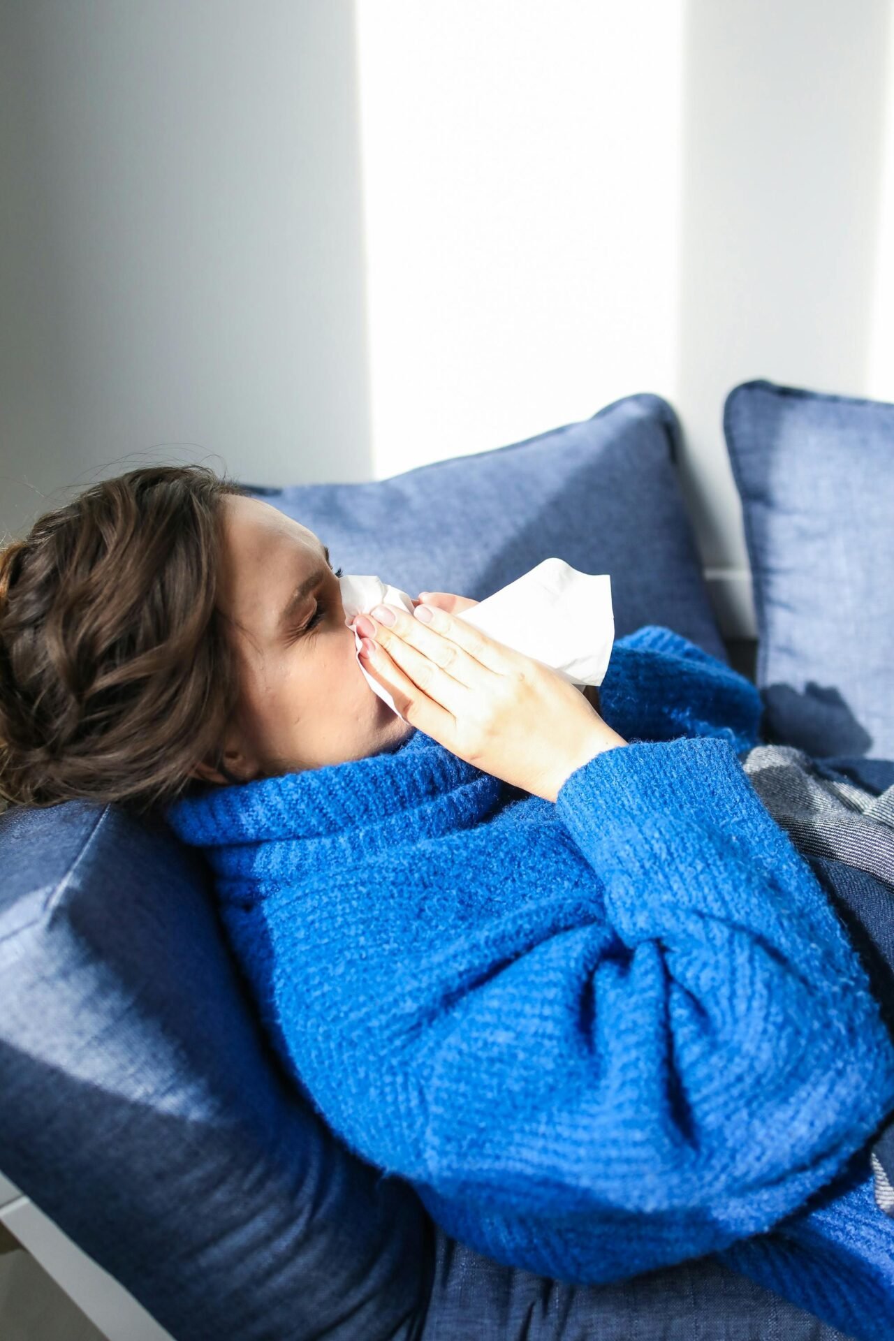 Frau im blauen Pullover liegt auf einem Sofa und putzt sich die Nase, um Glück und Gesundheit zu wünschen.