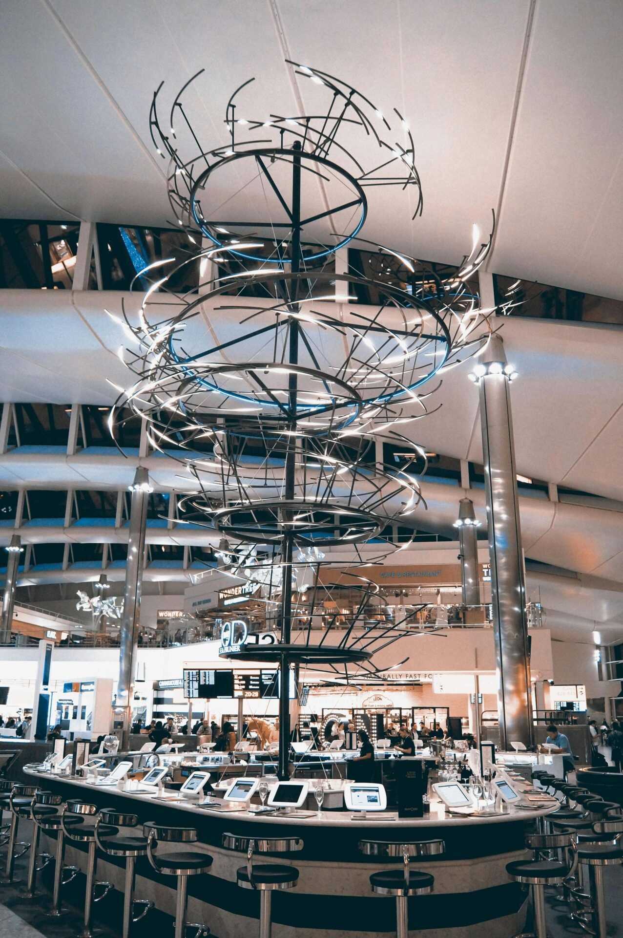 Moderne Londoner Flughafenbar mit einer spiralförmigen Metallskulptur und digitalen Bildschirmen unter einer hohen, gewölbten Decke.