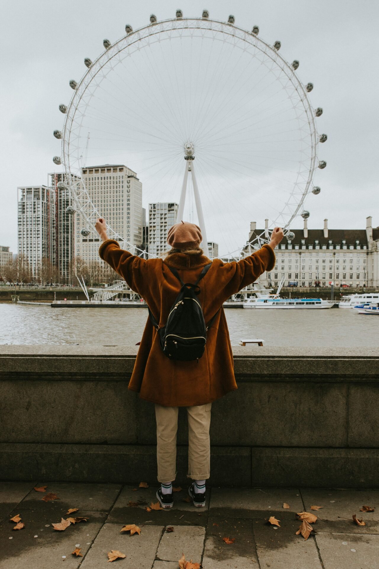 Eine Person in einem braunen Mantel steht vor dem London Eye in London, die Arme am Fluss erhoben.