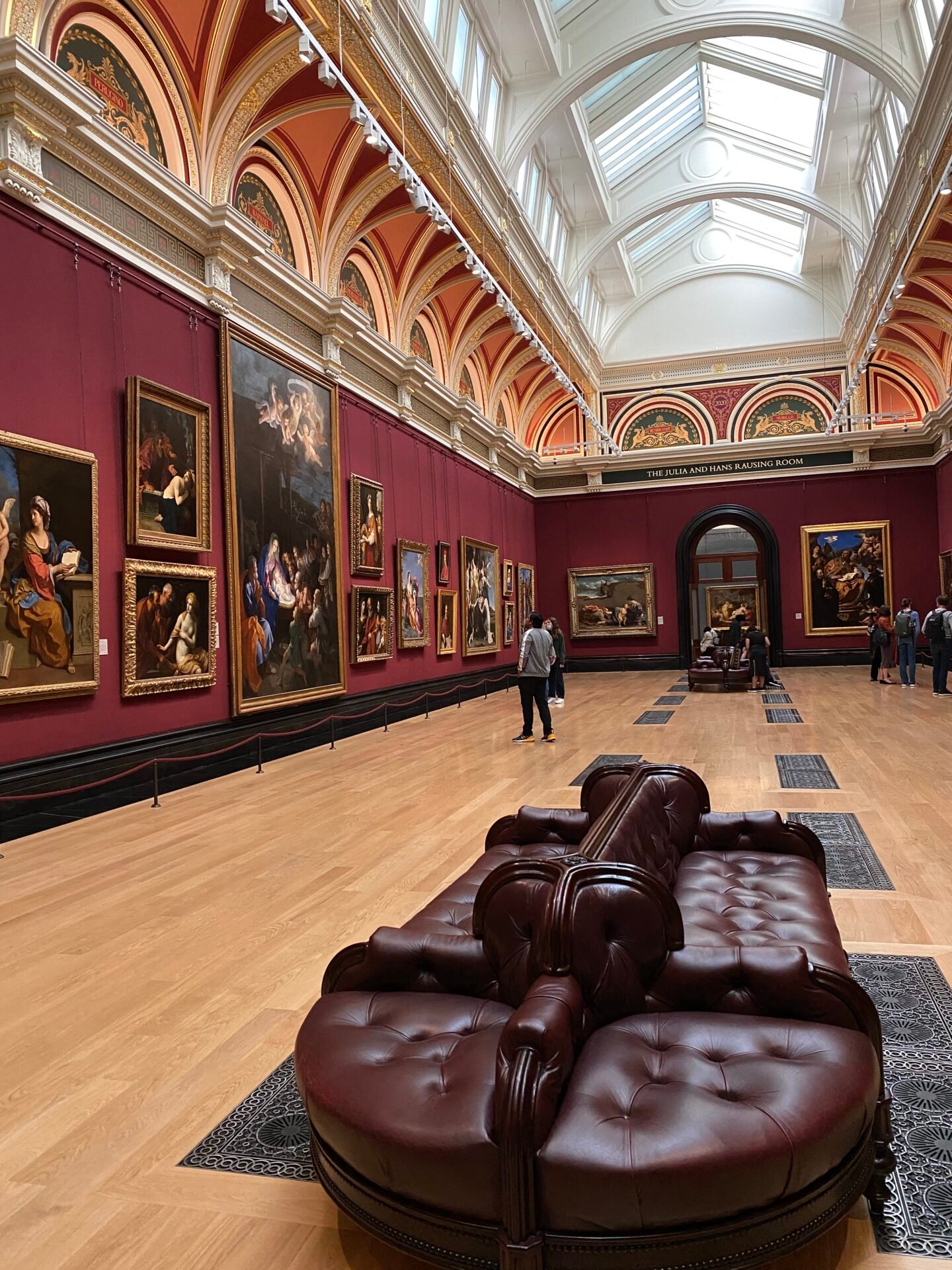 Eine große Londoner Kunstgalerie mit roten Wänden, großen Gemälden und Plüschsitzen.