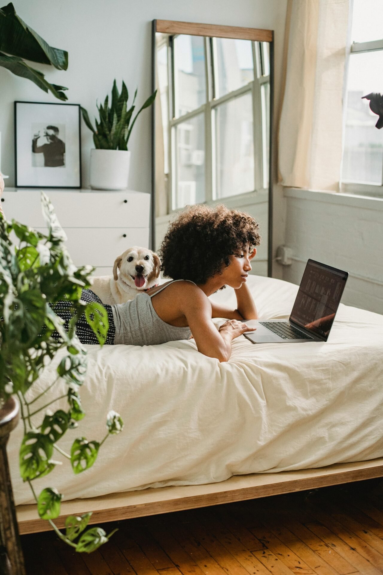 Frau nimmt einen Mentalitätswechsel vor, während sie ihren Laptop benutzt, ihr Hund an ihrer Seite in einem hellen Schlafzimmer.