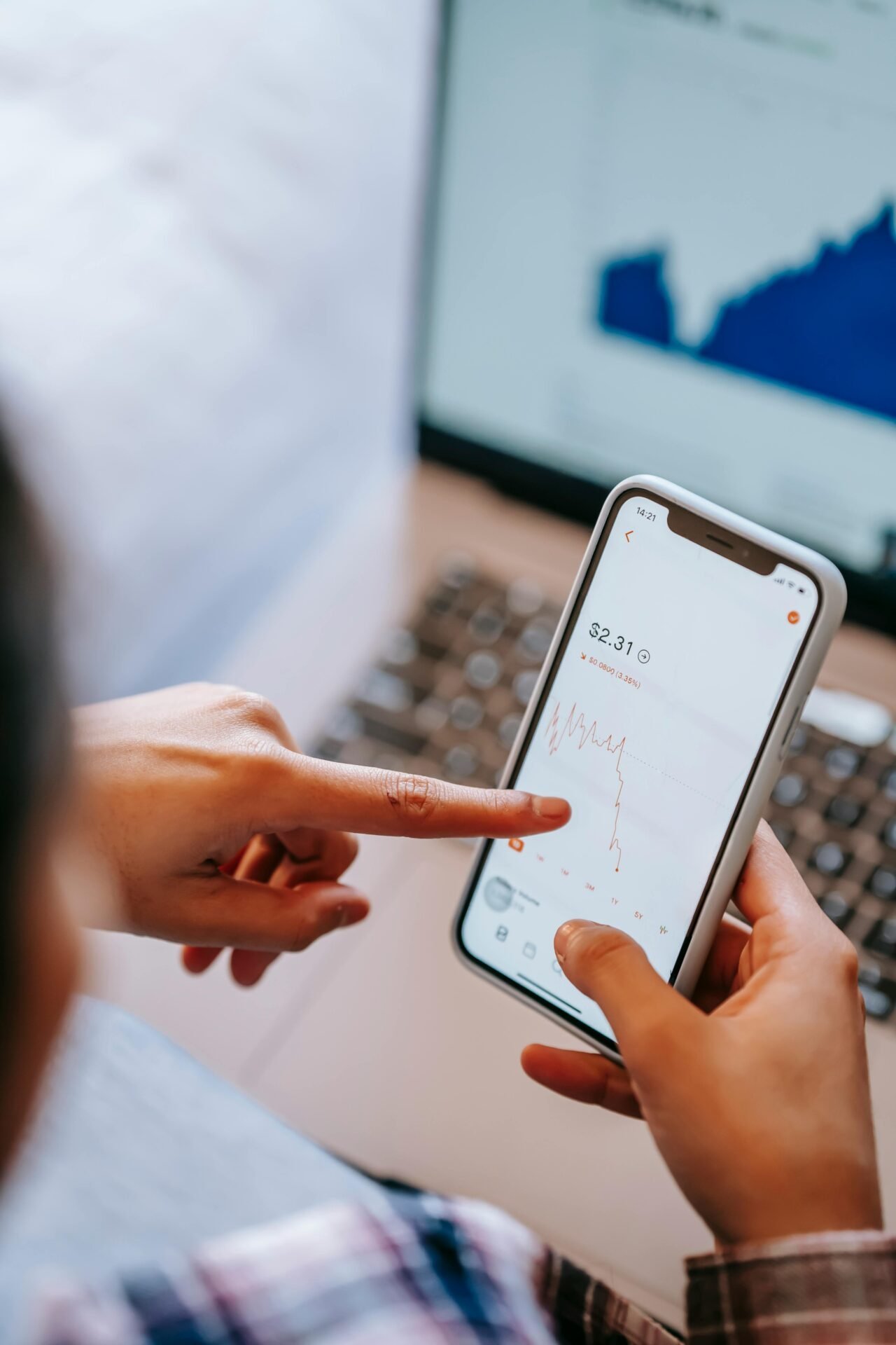 Person analysiert fallende Aktienkurse auf Smartphone, mit Online-Geschäftsdiagramm auf Laptop in der Nähe.