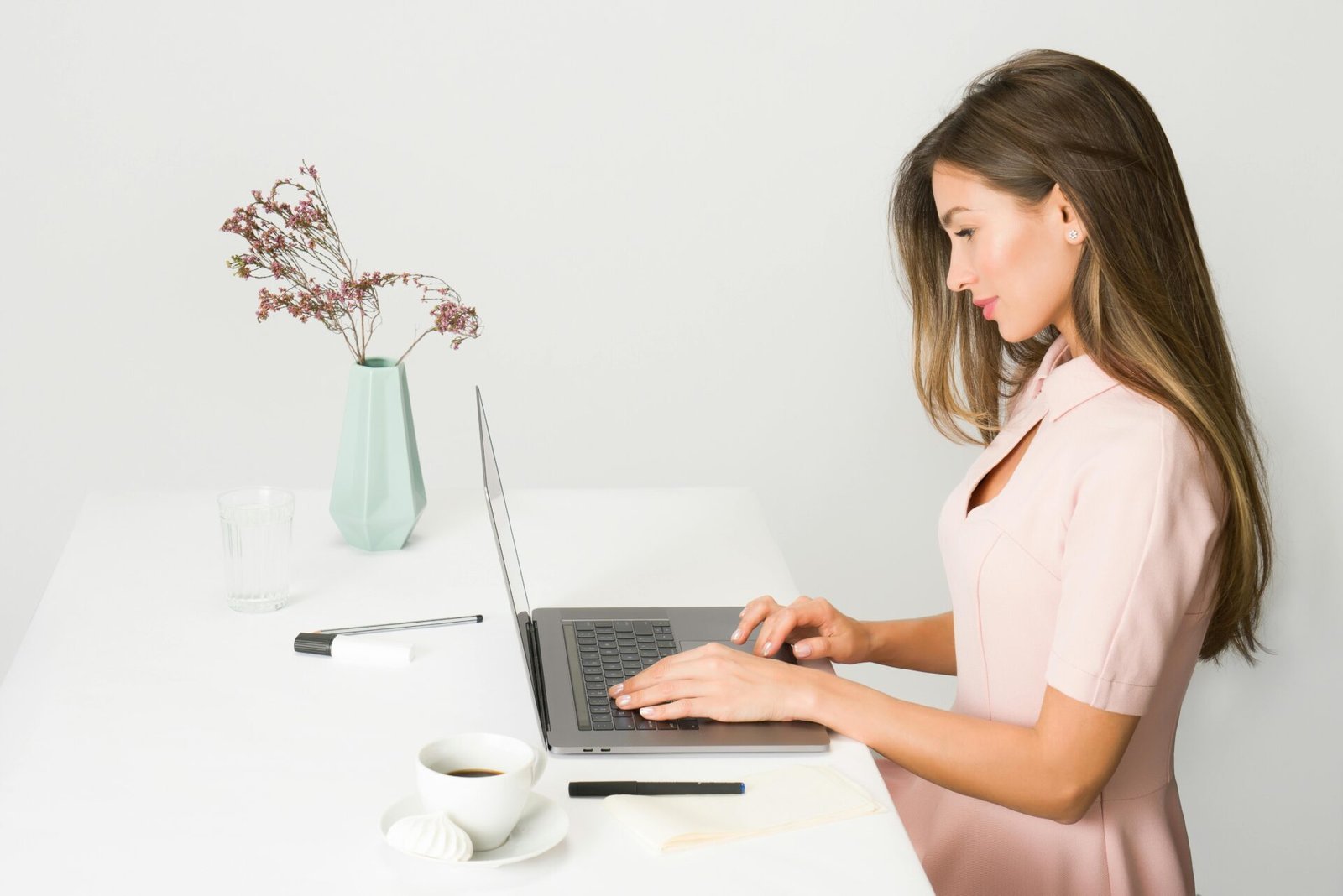 Frau im rosa Kleid betreibt ihr Online-Geschäft an einem weißen Schreibtisch mit Kaffee und einer Vase.