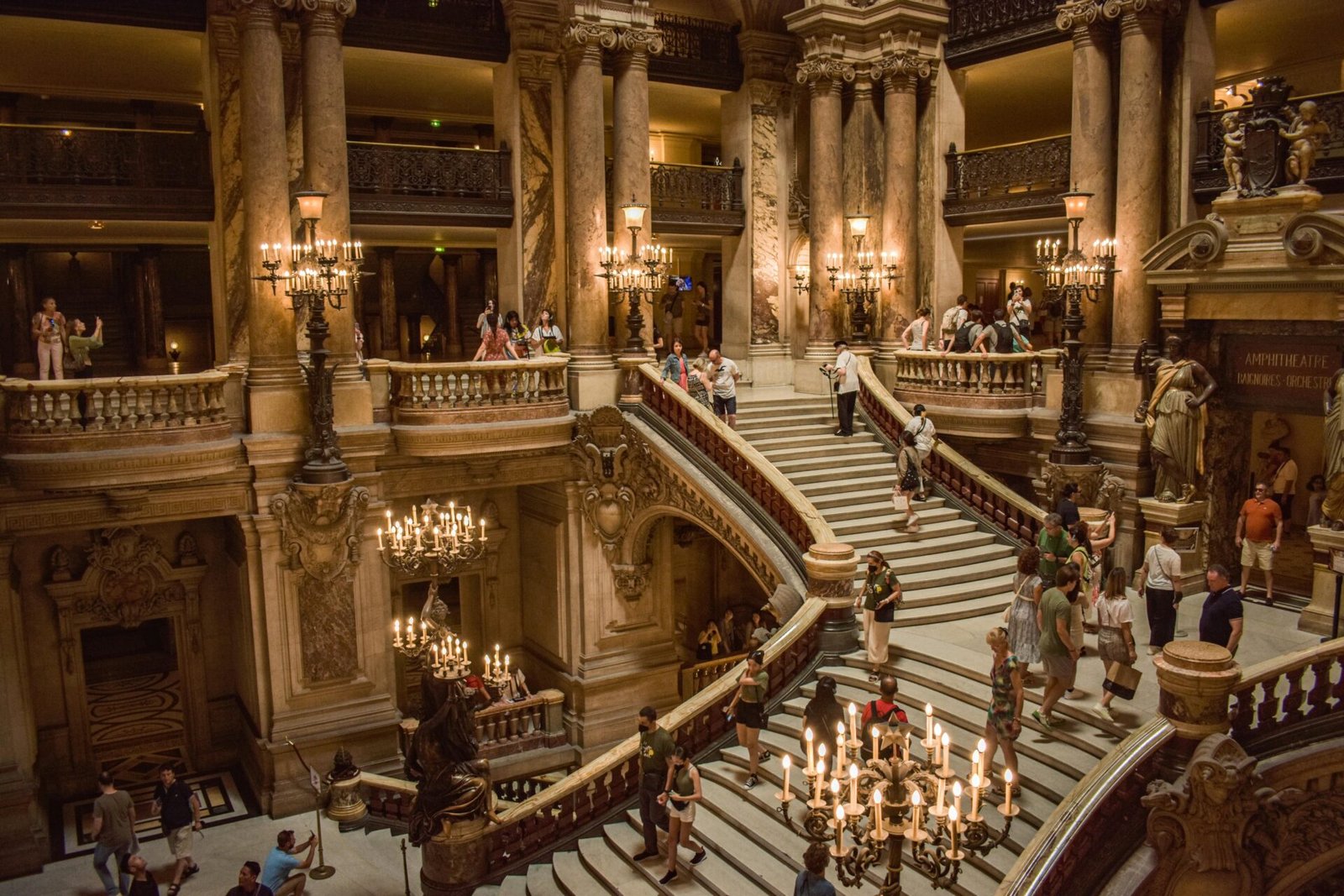 Großes Treppenhaus in einem historischen Pariser Gebäude mit Menschen, die gehen und Kronleuchtern, die leuchten.