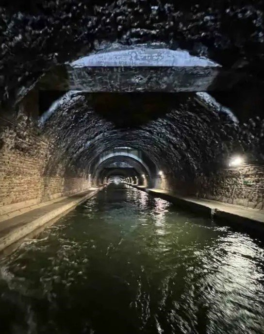 Ein schwach beleuchteter Pariser Tunnel, durch dessen Mitte Wasser fließt, mit gewölbten Steinwänden.