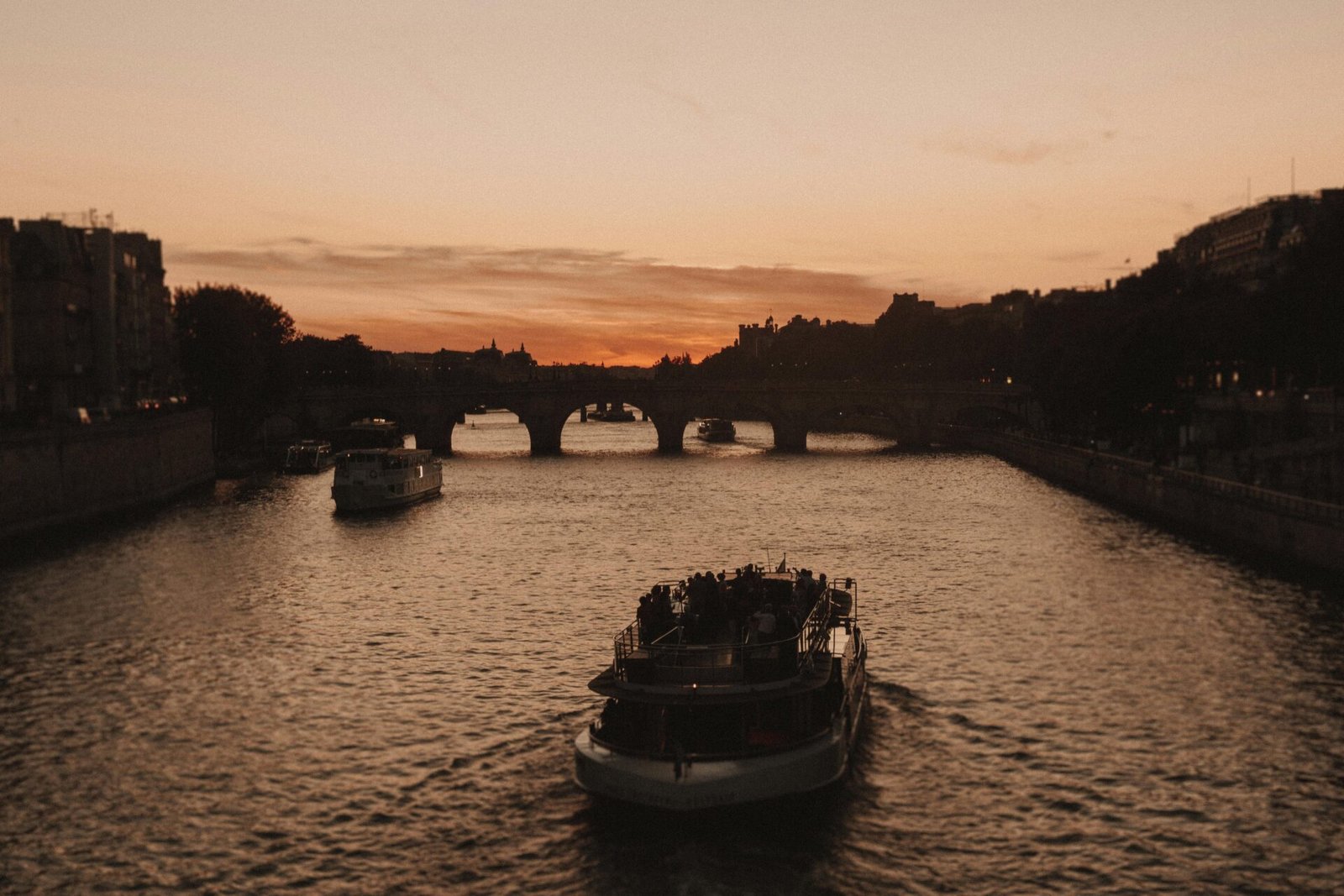 Ein Boot fährt bei Sonnenuntergang auf einem Pariser Fluss mit einer Steinbrücke im Hintergrund.
