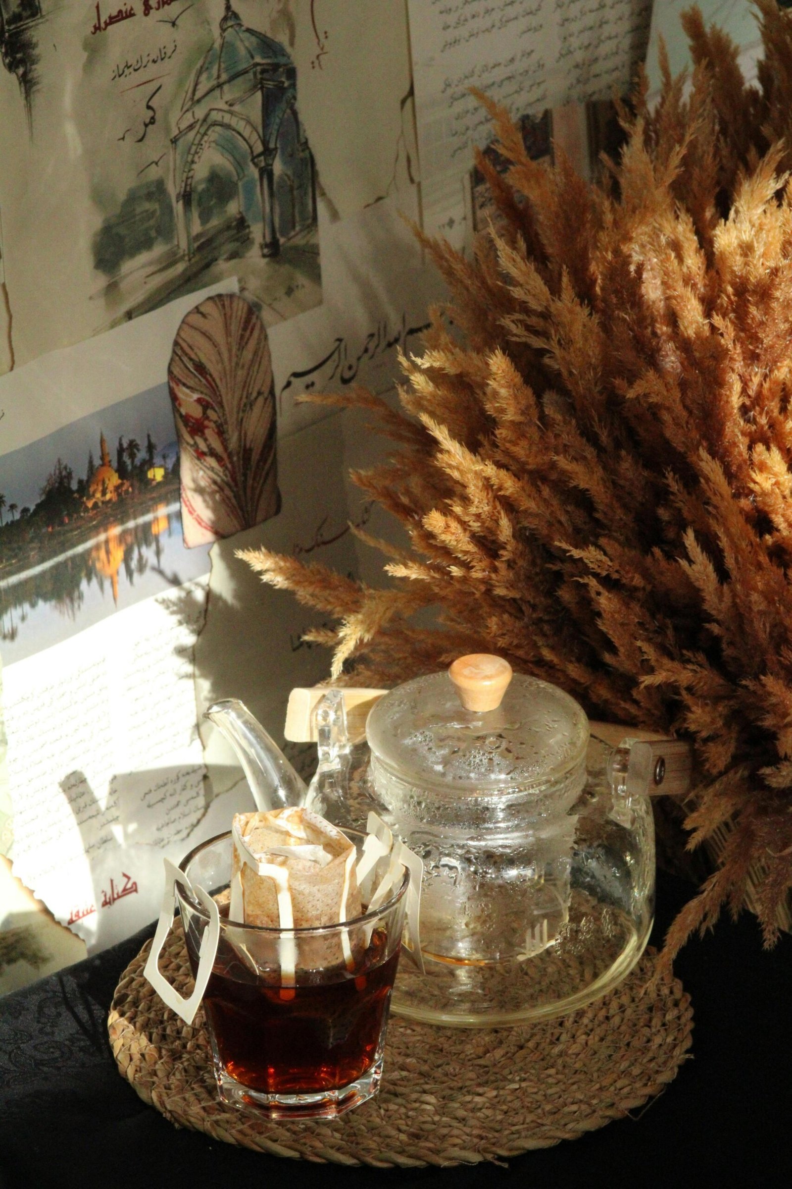 Eine gläserne Teekanne und eine Teetasse vor einem Hintergrund aus getrockneten Ähren und Fotos an der Wand symbolisieren die Kraft kleiner Rituale im Alltag.