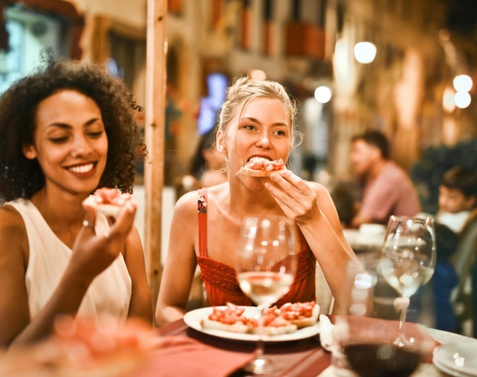 Zwei Frauen sitzen an einem Restauranttisch im Freien und genießen ihr Essen. Die eine lächelt und hält eine Scheibe Brot in der Hand, die andere beißt hinein. Der Tisch ist mit Weingläsern gedeckt, und der Hintergrund ist leicht verschwommen, was eine warme Atmosphäre suggeriert.