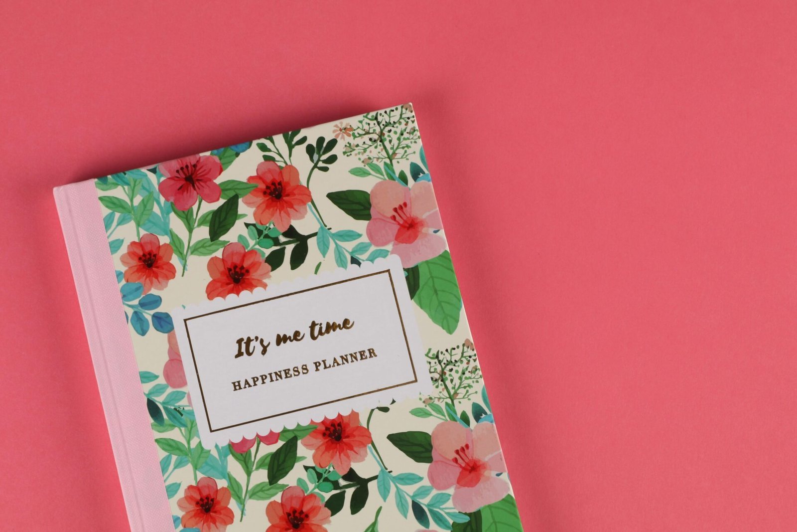 Ein Notizbuch mit Blumenmotiv und dem Titel „It's me time: Happiness Planner“ liegt auf einem korallenfarbenen Hintergrund. Der Einband ist mit leuchtenden Blumen und grünen Blättern verziert.