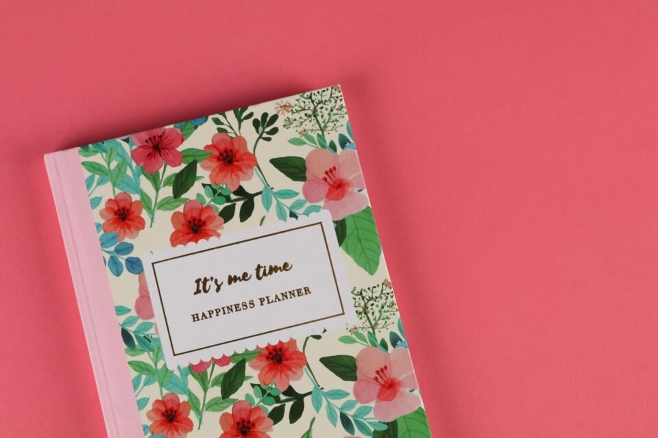 Ein Notizbuch mit Blumenmotiv und dem Titel „It's me time: Happiness Planner“ liegt auf einem korallenfarbenen Hintergrund. Der Einband ist mit leuchtenden Blumen und grünen Blättern verziert.