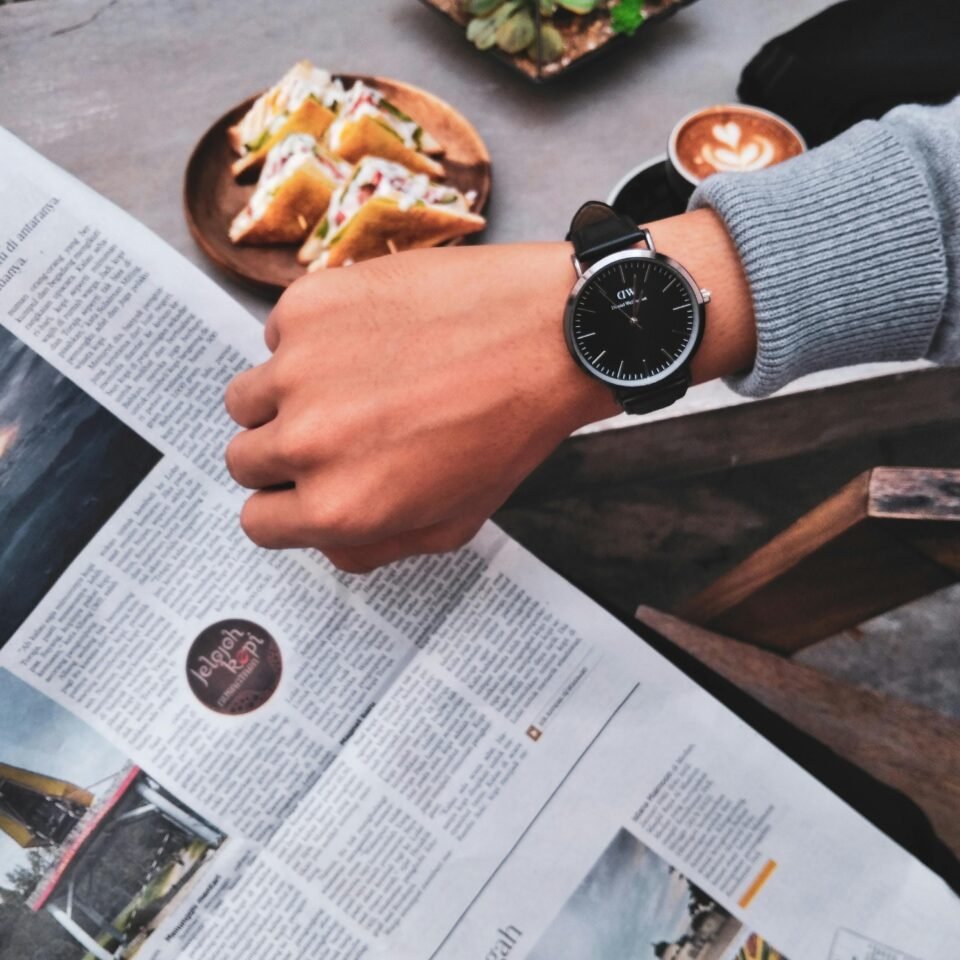 Eine Person trägt eine schwarze Uhr und schaut auf die Uhrzeit, während sie eine Zeitung in der Hand hält. Auf dem Tisch stehen eine Tasse Kaffee und ein Teller mit Sandwiches. Die Person trägt einen grauen Pullover.