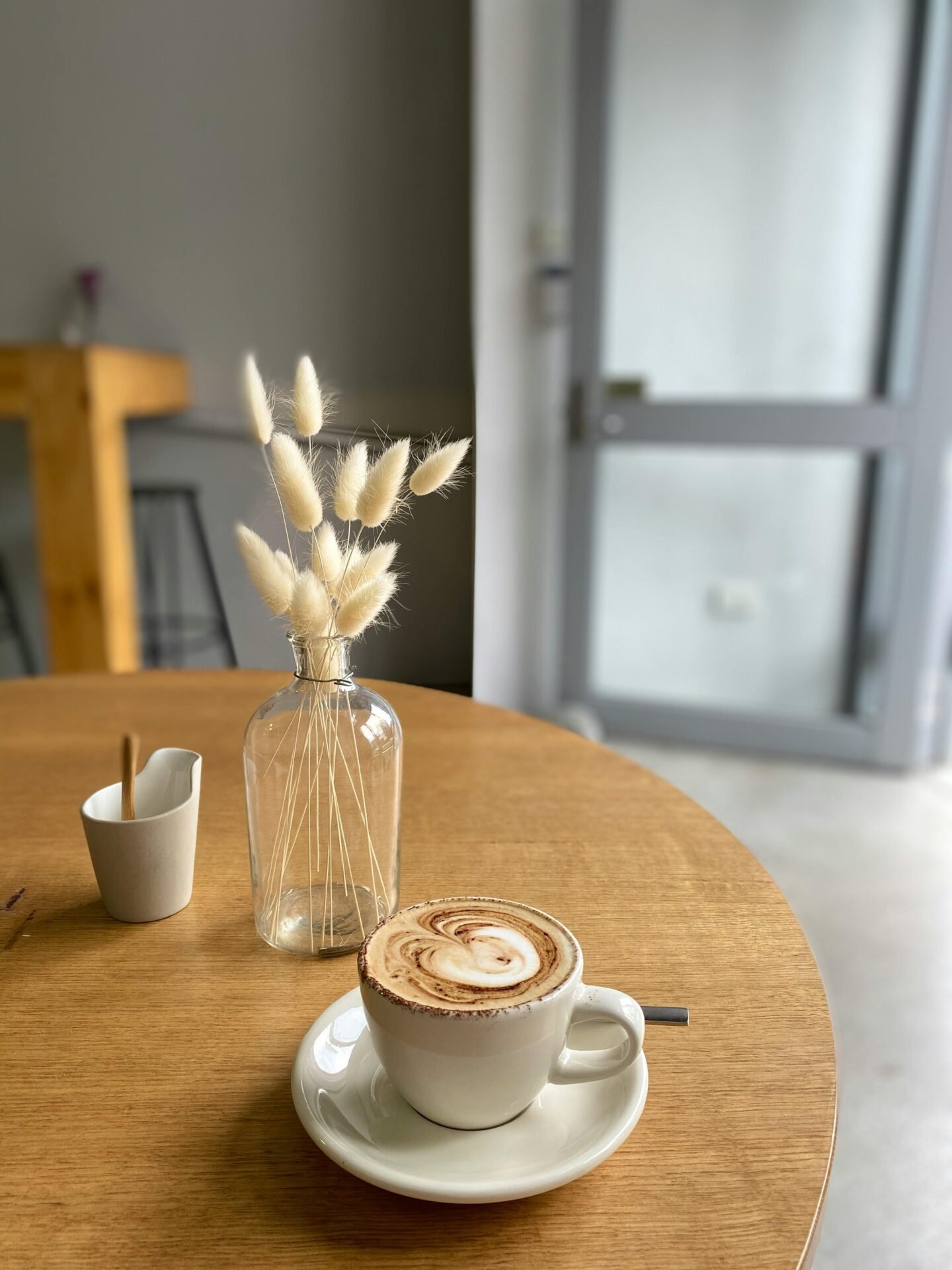 Ein Kaffee steht auf einem runden Holztisch mit einer Blumenvase. In der Vase sind getrocknete Gräser.