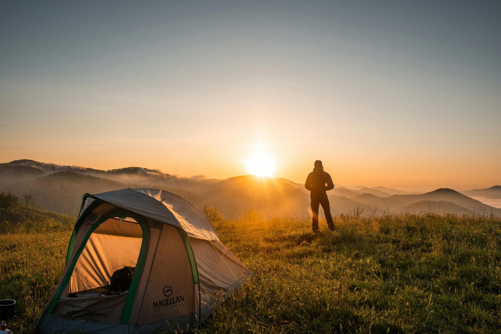 Eine Person steht neben einem Zelt auf einem grasbewachsenen Hügel und beobachtet den Sonnenaufgang über den fernen, nebligen Bergen. Der Himmel ist klar und in warmen Orange- und Gelbtönen gehalten. Die Szene vermittelt ein Gefühl von Ruhe und Abenteuer.