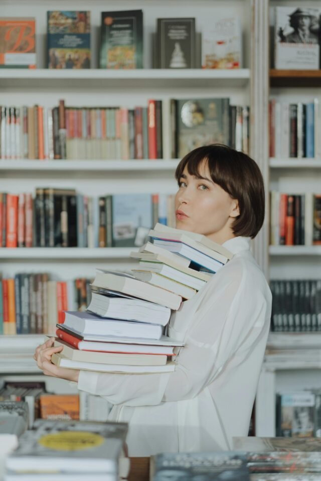 Glückselige Person mit einem hohen Stapel Bücher in einem gemütlichen Buchladen.