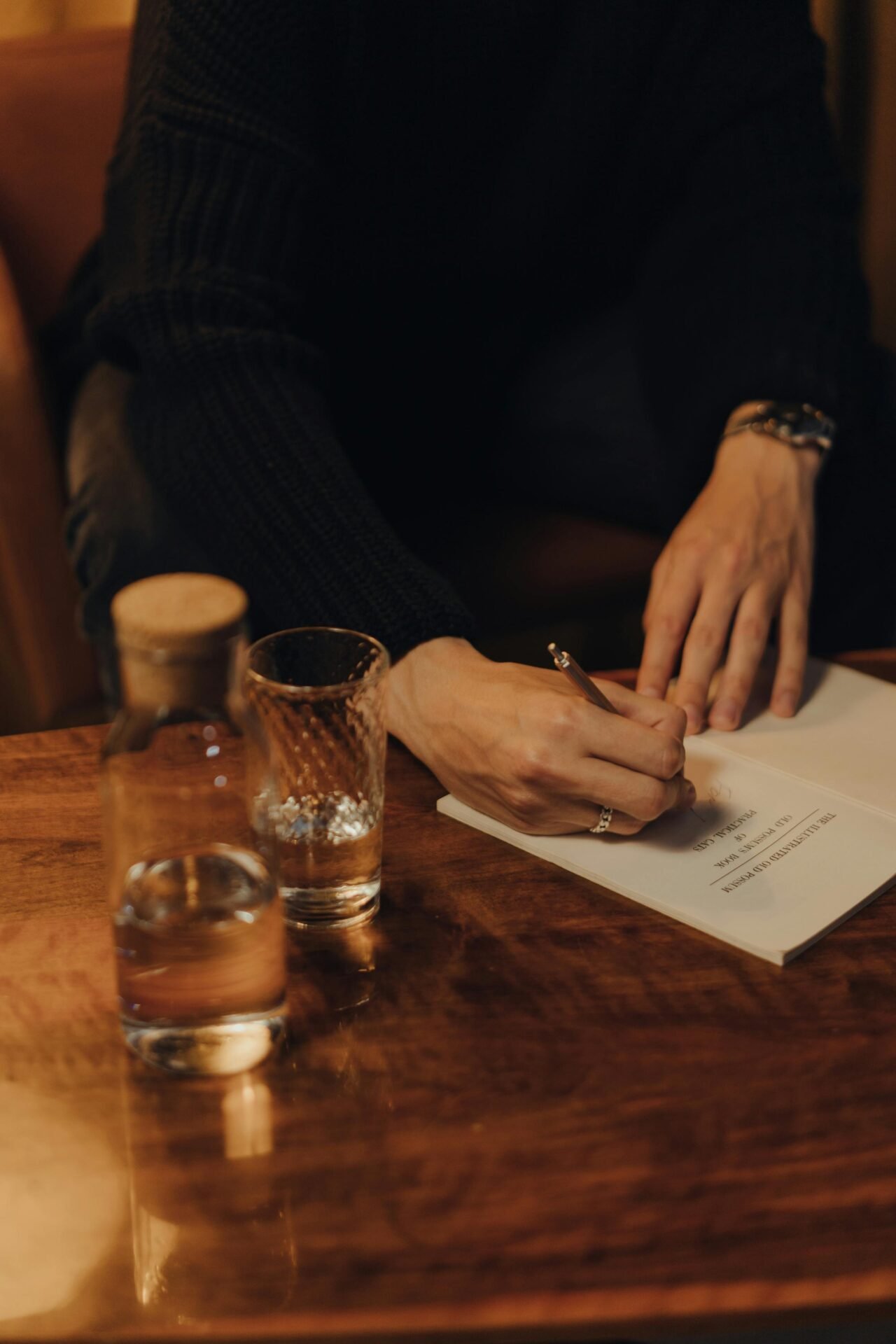 Eine Person schreibt an einem Tisch mit einem Glas Wasser und einer Wasserflasche in ein Journal.