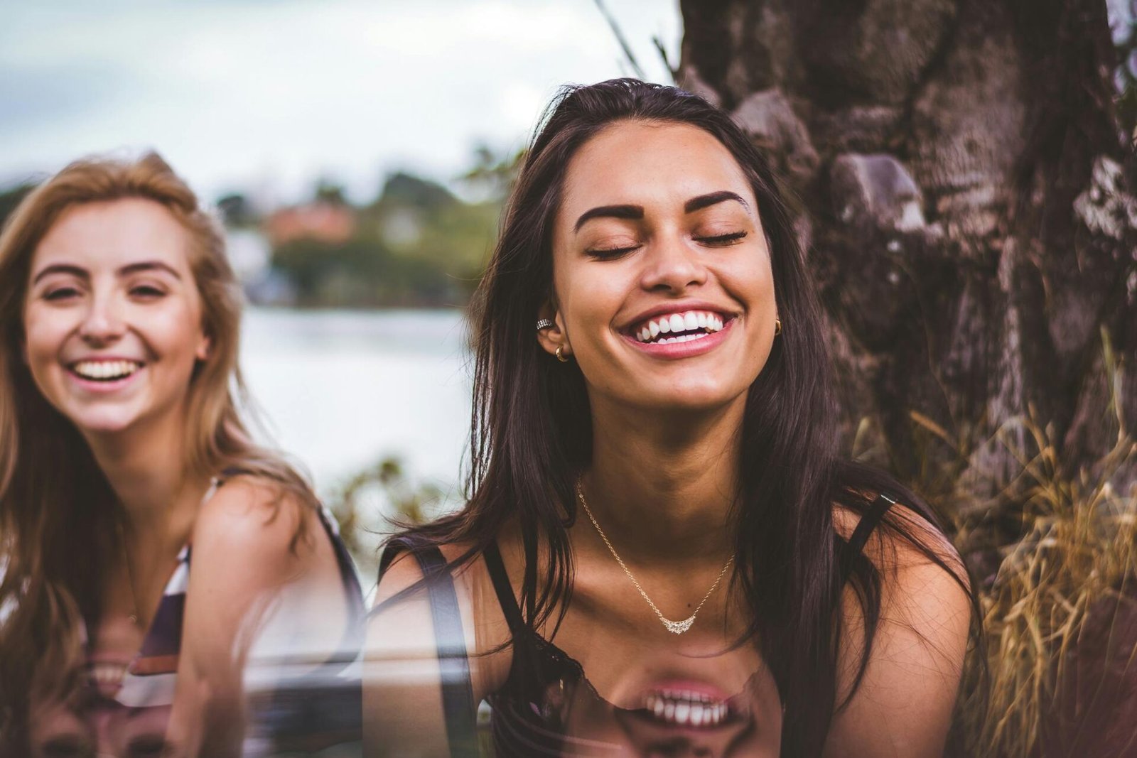 Zwei Frauen lachen im Freien neben einem Baum, vor einem unscharfen Hintergrund aus Grün und Wasser. Die eine hat langes dunkles Haar, die andere helles. Beide tragen lässige Oberteile und wirken fröhlich.