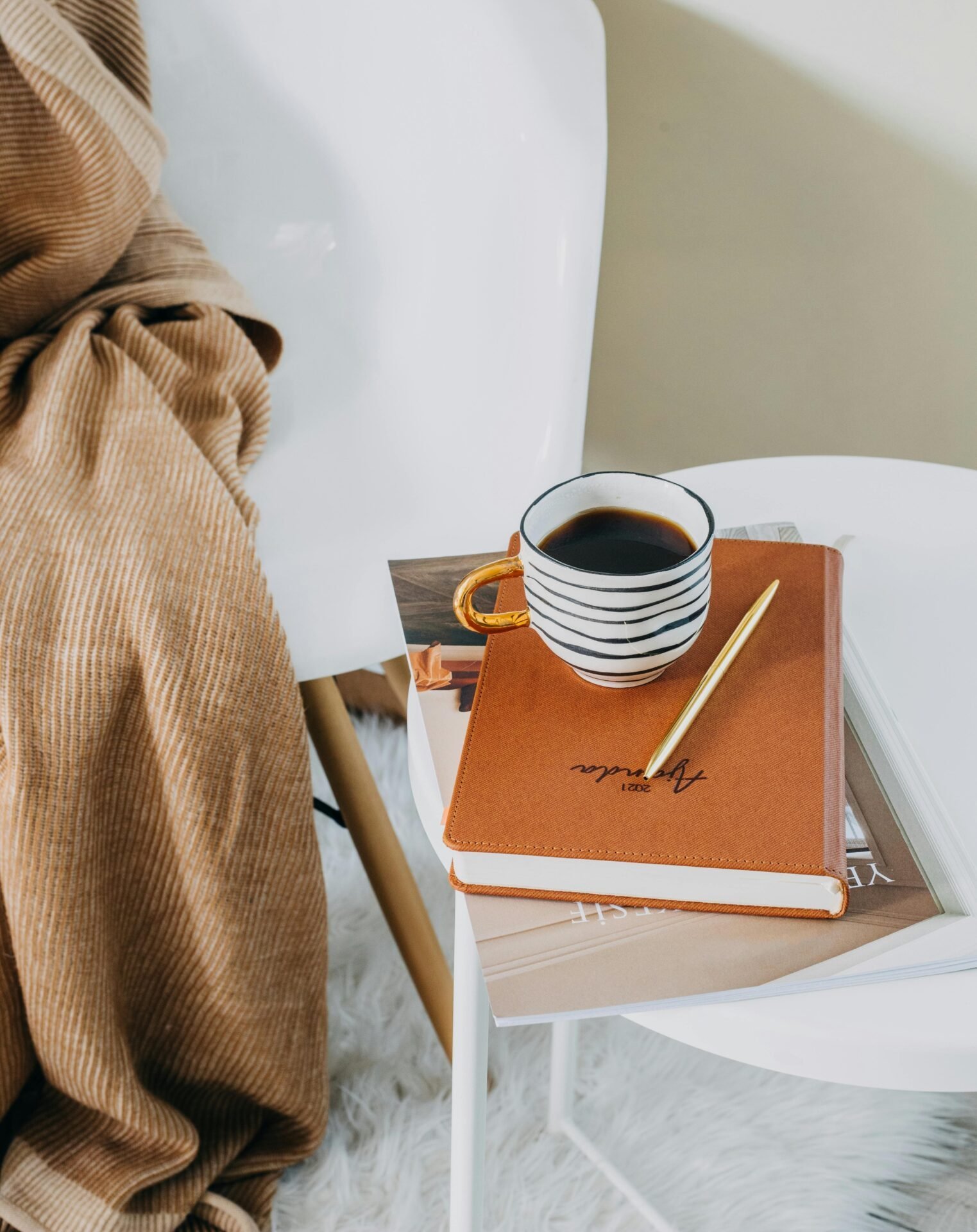 Ein Buch zur Jahres-Organisation liegt neben jemandem auf einem Beistelltisch mit einem goldenen Kugelschreiber und einer gestreiften Tasse Kaffee.