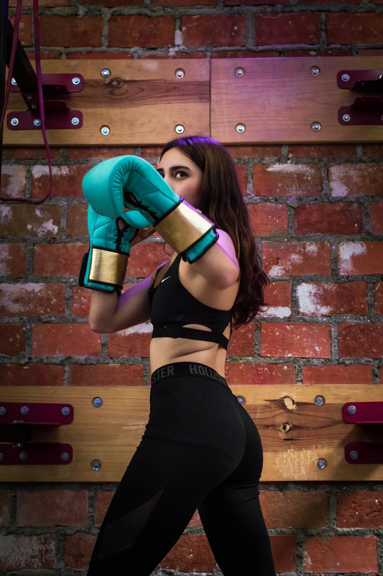 Eine Frau in schwarzem Sportoutfit steht in Boxhaltung vor einer Backsteinmauer. Sie trägt blaue Boxhandschuhe und hat langes, wallendes Haar. Die Szenerie besteht aus Holzpaneelen und einer warmen Lichtstimmung.