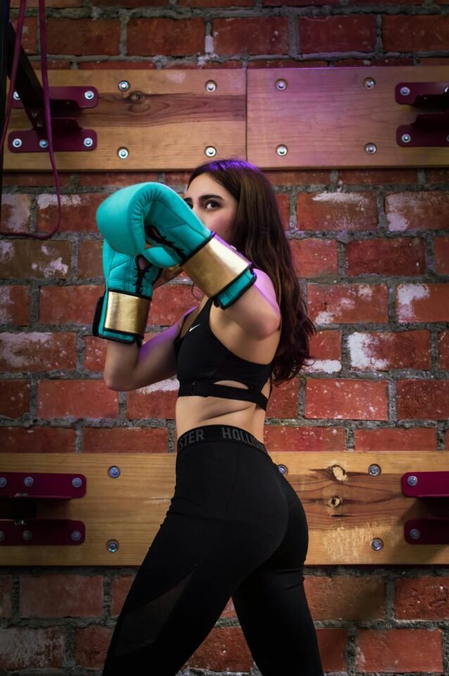 Eine Frau in schwarzem Sportoutfit steht in Boxhaltung vor einer Backsteinmauer. Sie trägt blaue Boxhandschuhe und hat langes, wallendes Haar. Die Szenerie besteht aus Holzpaneelen und einer warmen Lichtstimmung.