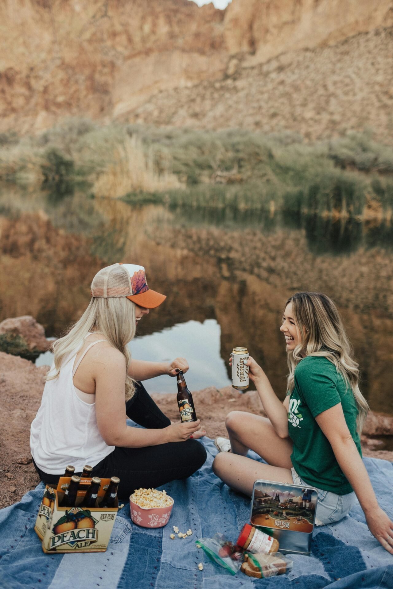 Zwei junge Frauen haben es hyggelig: Sie sitzen lachend auf einer Decke am Fluss mit Snacks wie Popcorn und Peach Ale und unterhalten sich.