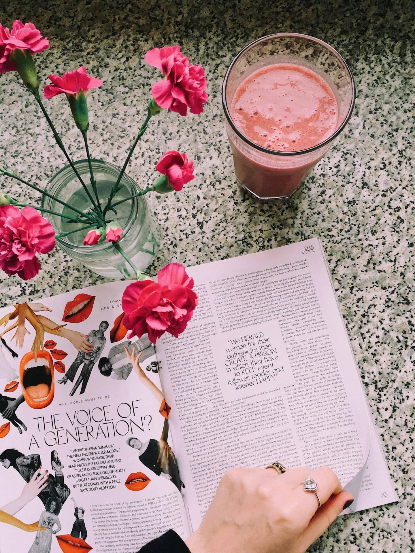 Eine Frau blättert in einem Magazin, um passende Bilder zu finden. Auf dem Tisch stehen ein Smoothie und pinke Blumen in einem Glas.