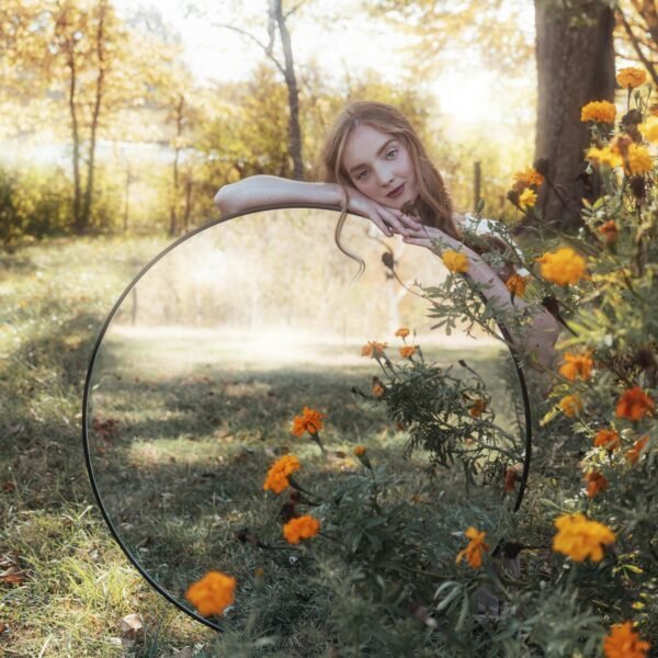 Eine Person mit langen Haaren lehnt an einem großen runden Spiegel, der im Gras zwischen orangefarbenen Blumen steht. Der Spiegel reflektiert den sonnigen, grünen Hintergrund. Die Szene ist ruhig und von der Natur inspiriert.