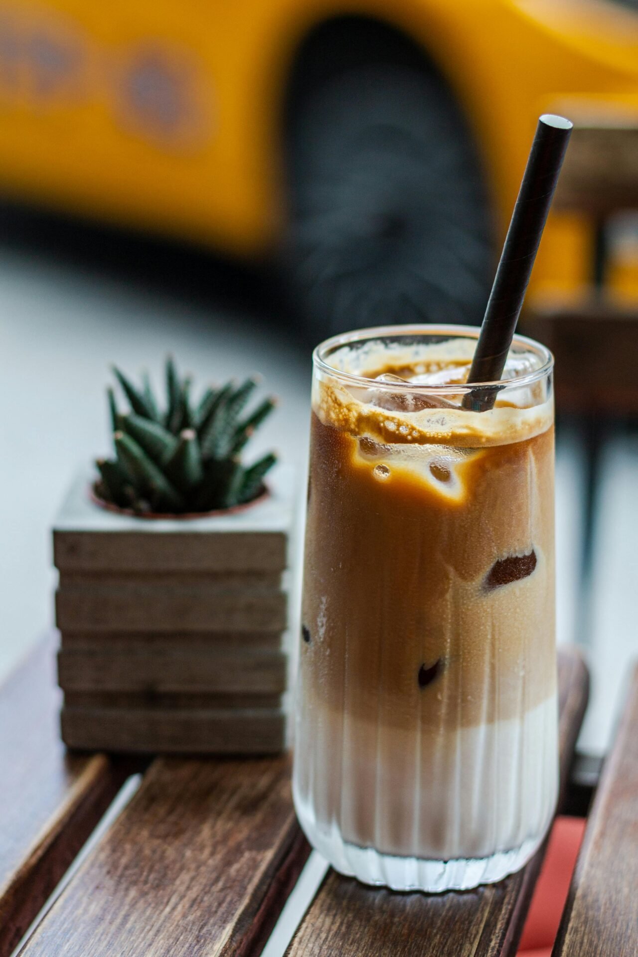Ein Glas Eiskaffee mit einem Stromhalm. Der Eiskaffee hat eine hellbraune Farbe und ist unten weiß. Im Hintergrund steht eine Sukkulente.