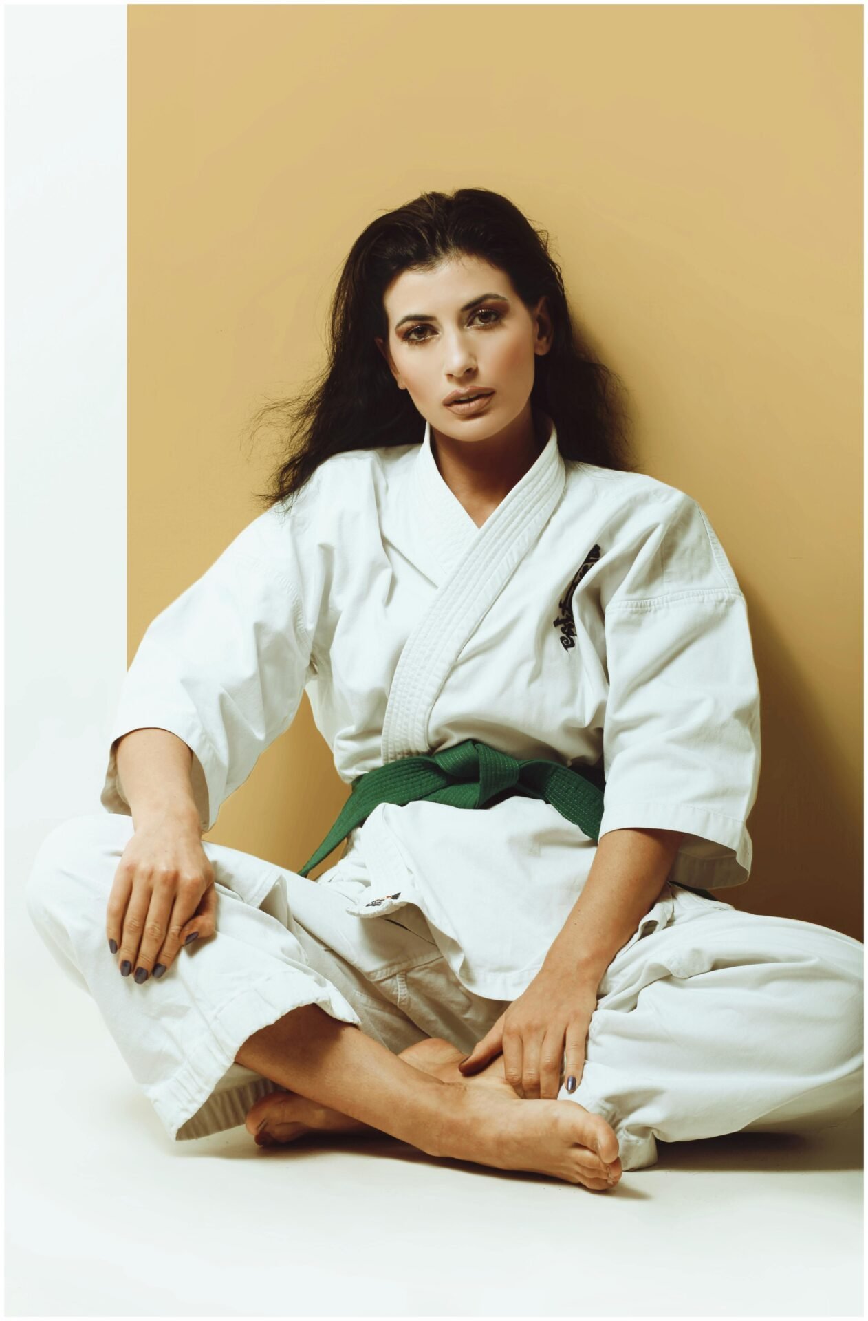 Eine dunkelhaarige Frau in Judo-Outfit mit grünem Gürtel sitzt im Schneidersitz an der Wand.