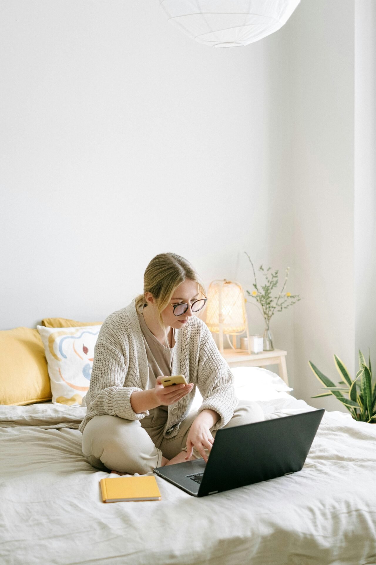 Eine blonde Frau mit Brille und Strickjacke sitzt auf ihrem Bett und organisiert etwas mit Handy und Laptop.