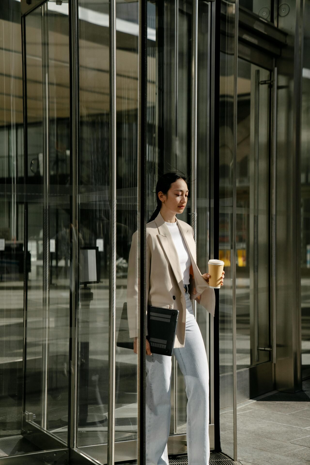 Frau im beigen Blazer kommt mit Kaffee in der Hand aus einer Glastür – ein gesünderer Lebensstil-Moment.