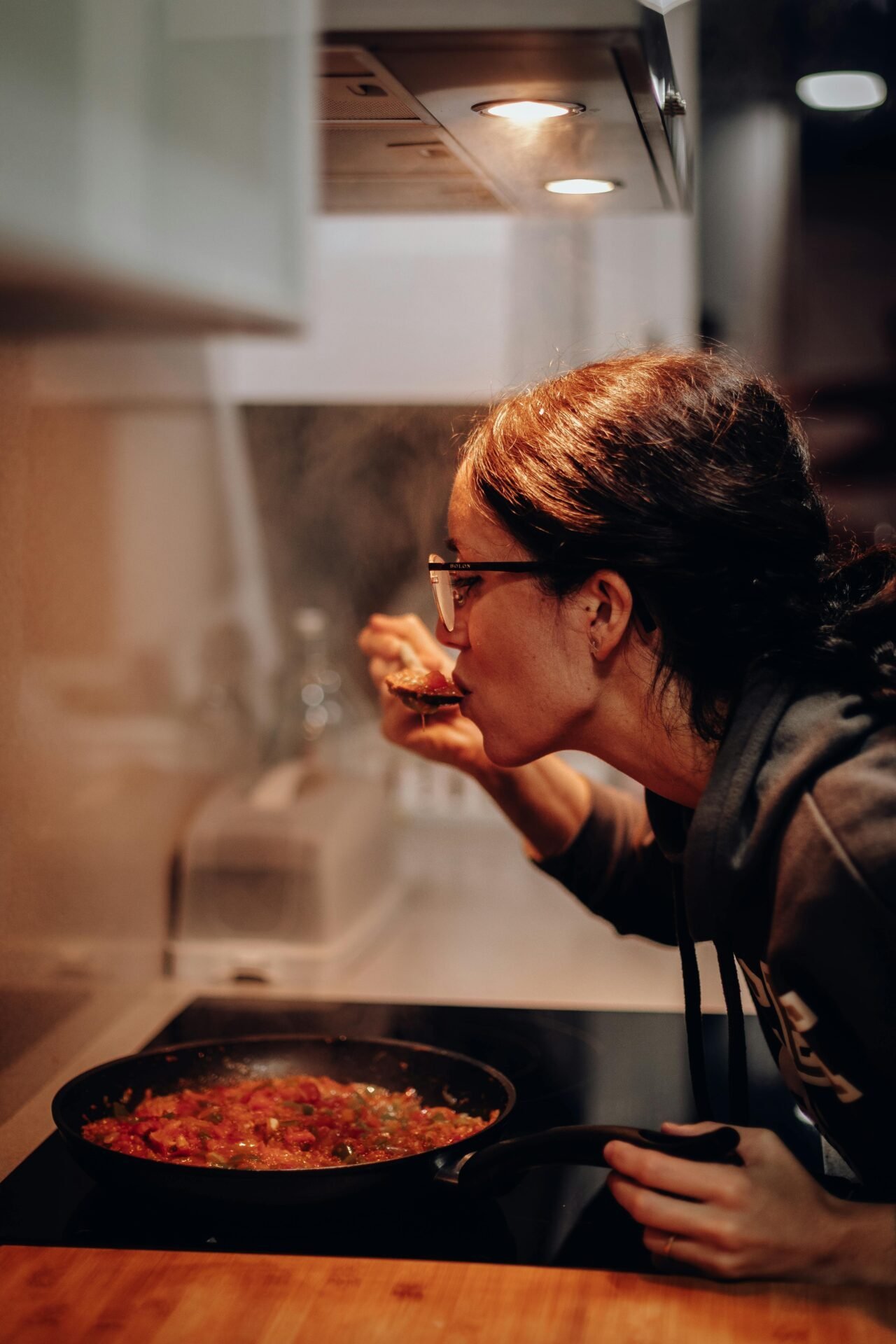 Eine fröhliche Frau beugt sich über einen Herd und probiert eine Tomatensoße vom Kochlöffel.