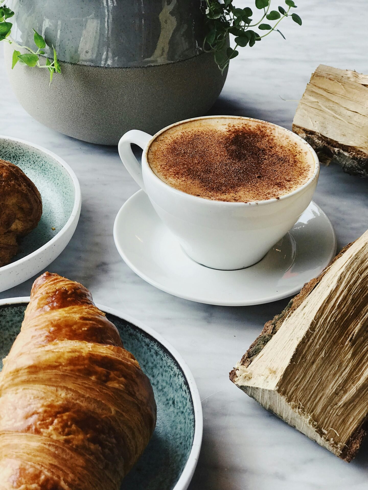 Eine Tasse Kaffee und ein Teller mit einem Croissant. Im Hintergrund steht eine Topfpflanze.